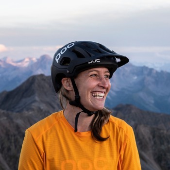 Portrait of Monika Freiburghaus from BSI Software wearing a helmet and orange shirt, with alpine mountains in the background