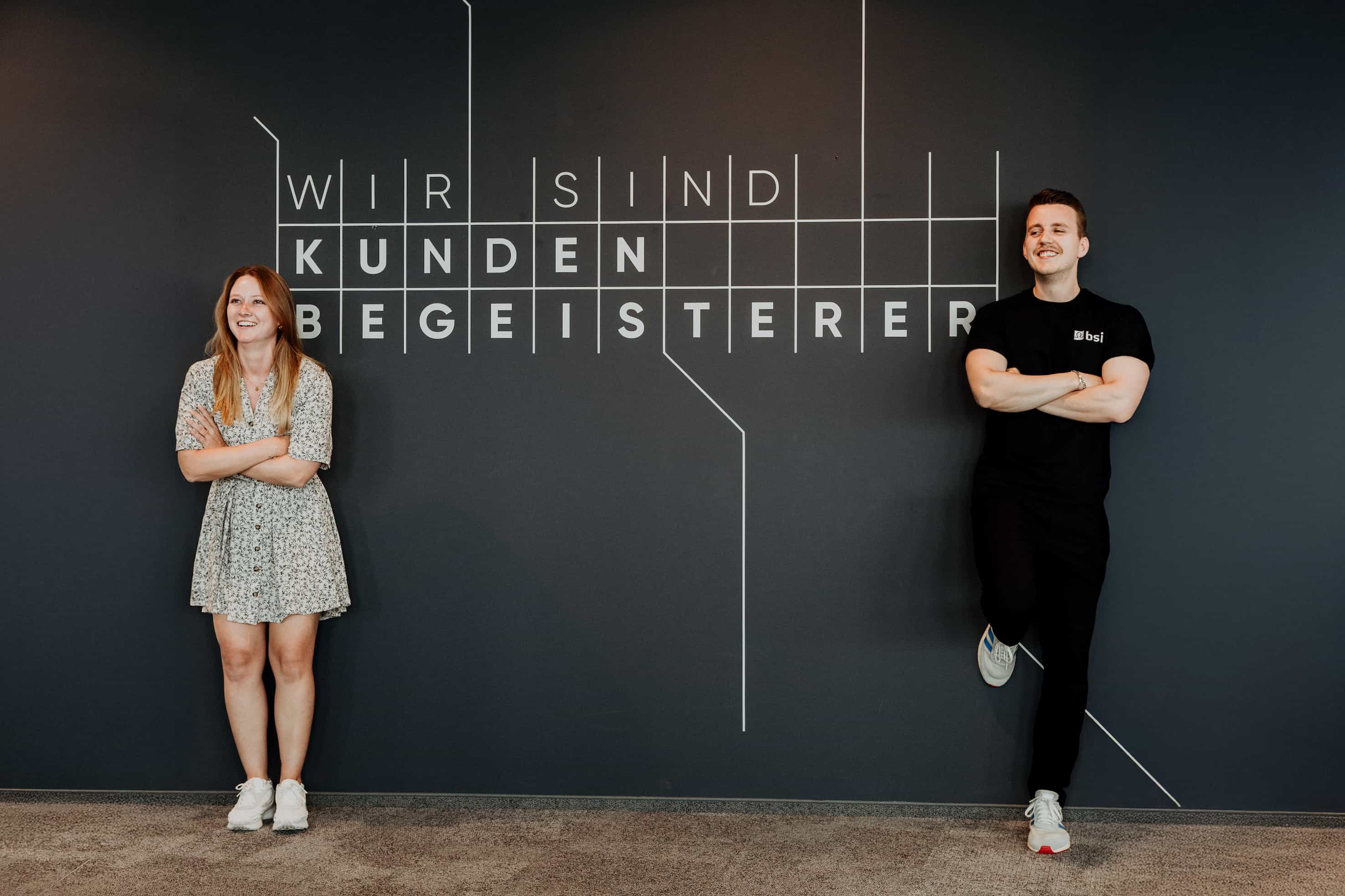 Zwei Mitarbeitende stehen lächelnd vor einer dunkelgrauen Wand mit der Aufschrift „Wir sind Kundenbegeisterer“. Links steht eine Frau in einem hellen geblümten Kleid, rechts ein Mann in schwarzer Kleidung mit verschränkten Armen; modernes Büroambiente und freundliche Teamatmosphäre.