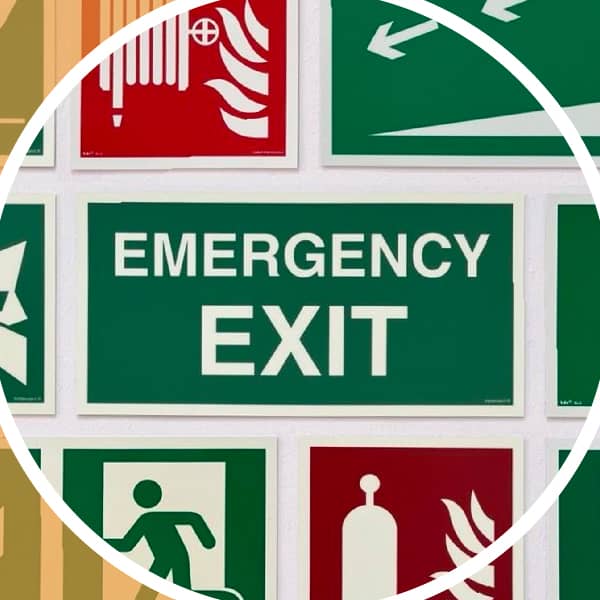Sammlung von Sicherheitsschildern, darunter ein großes grünes Schild mit der Aufschrift «Emergency Exit», umgeben von Piktogrammen für Fluchtwege, Feuerlöscher und Notausgänge.