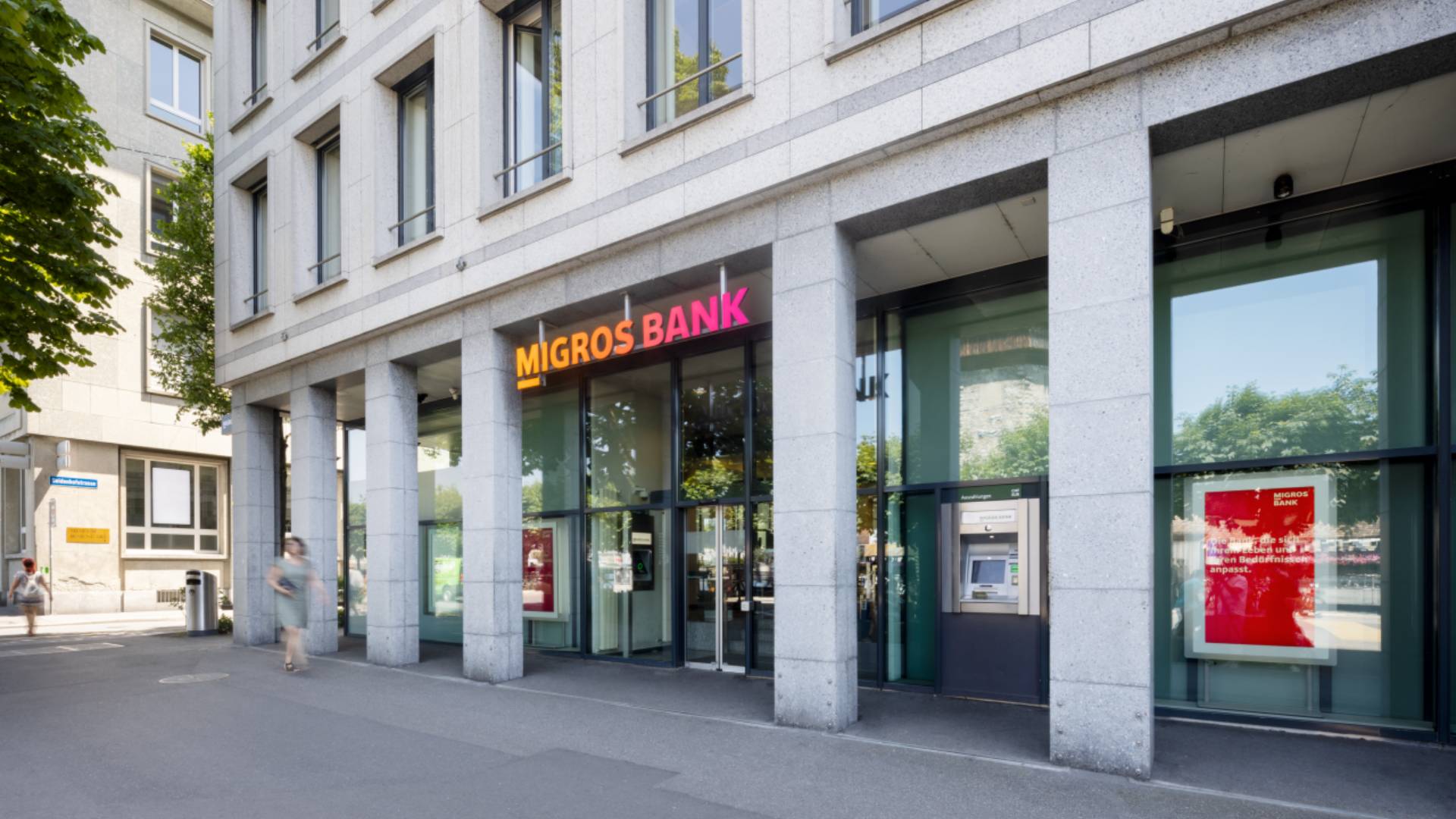 Vue extérieure d’une agence Migros Bank avec façade moderne en pierre, grandes baies vitrées et logo lumineux; passants devant l’entrée et bancomat visible, symbole de banque, d’expérience client et de customer relationship management avec la BSI Customer Suite