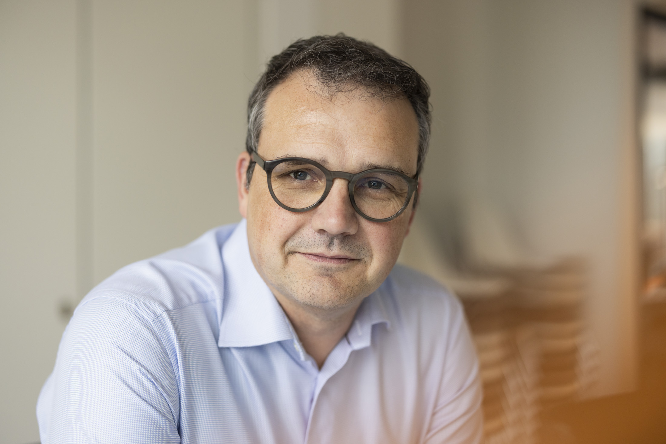 Porträt eines Mannes mit Brille und hellblauem Hemd, sitzend in einem modernen Büro mit unscharfem Hintergrund.
