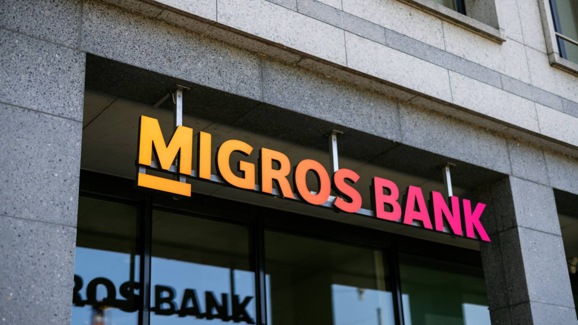 Vue extérieure d’une agence Migros Bank avec logo lumineux sur une façade moderne; symbole de banque, d’expérience client et de customer relationship management avec la BSI Customer Suite