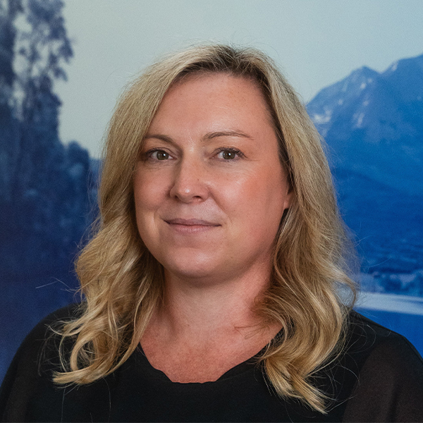 Porträt einer lächelnden Frau mit blonden, welligen Haaren vor einem Hintergrund mit Bergen und Waldlandschaft.