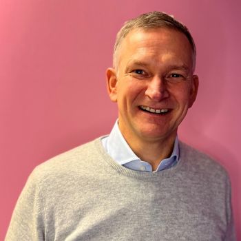 Porträtfoto eines Mannes im grauen Pullover mit blauem Hemd, stehend vor einer pinkfarbenen Wand und freundlich in die Kamera lächelnd.