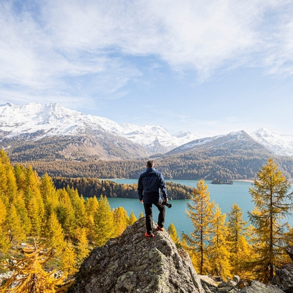 Urs Frick from BSI Software standing on a rock above Lake Sils in the Engadin, holding a camera in his right hand – surrounded by golden larch trees and snow-covered mountains.