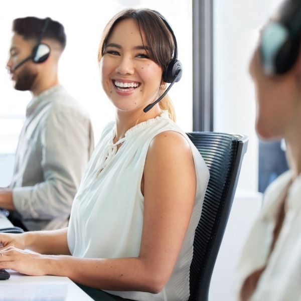Freundliche Callcenter-Mitarbeiterin mit Headset lächelt in die Kamera, während sie mit Kollegen im modernen Büro telefonischen Kundenservice anbietet.