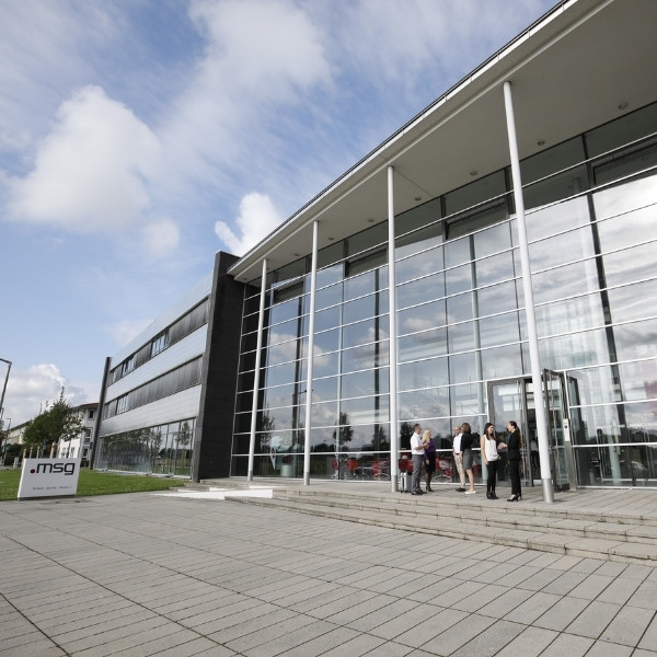 Exterior view of msg headquarters – a BSI Software partner – with a modern glass façade and people talking in front of the entrance.