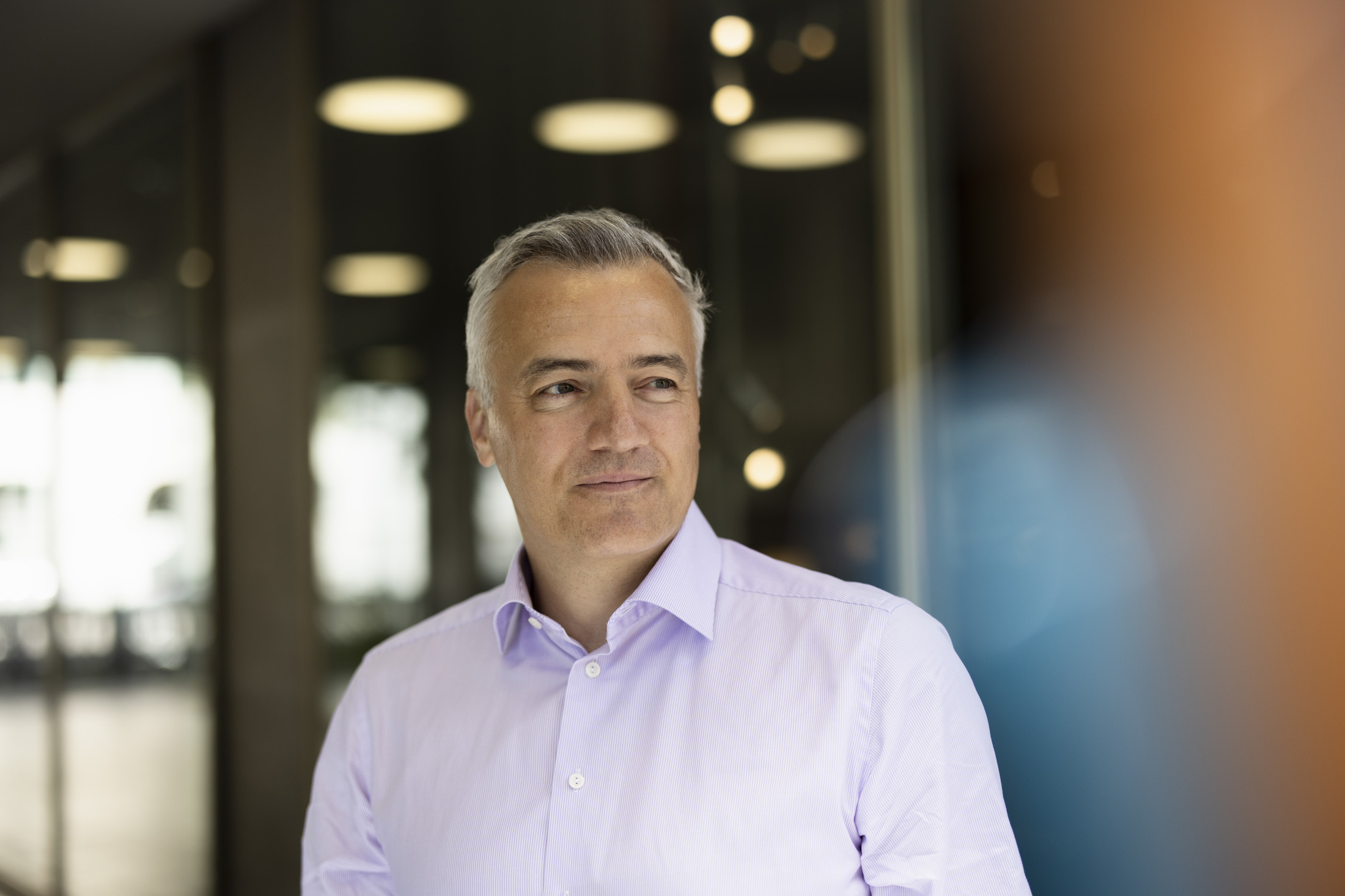 Porträt eines Mannes mit grauen Haaren im hellvioletten Hemd, stehend in einem modernen Innenraum und zur Seite blickend.