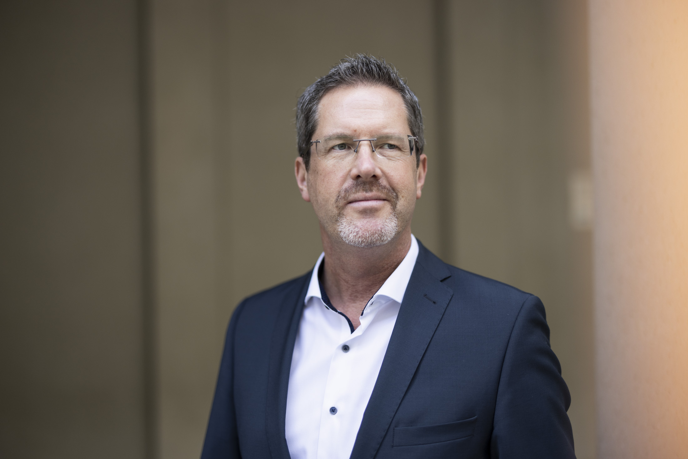 Porträt eines Mannes mit Brille im dunkelblauen Anzug und weißem Hemd, stehend vor neutralem Hintergrund und leicht zur Seite blickend.