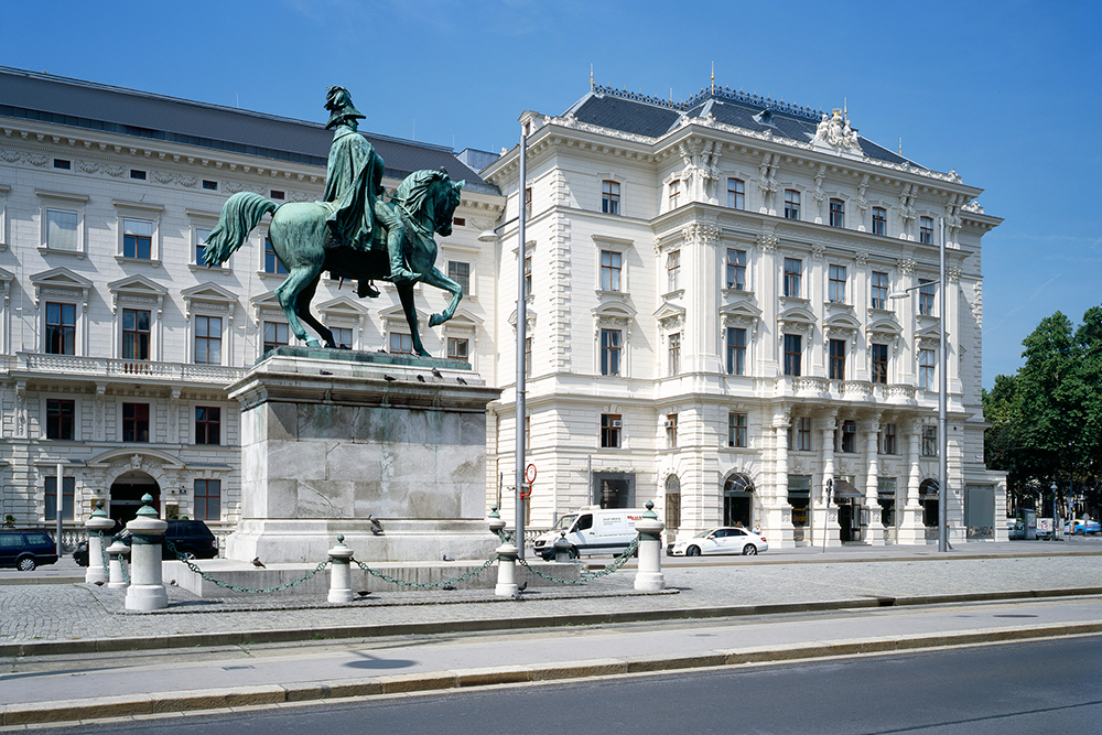 Historisches Reiterdenkmal vor einem prunkvollen Gründerzeitgebäude in Wien bei blauem Himmel.