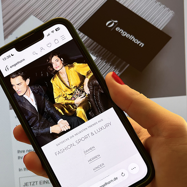 Smartphone in a woman's hand displaying the fashion retailer engelhorn's mobile app with the slogan “Discover the latest trends in Fashion, Sport & Luxury” and an image of stylishly dressed models; a blurred engelhorn loyalty card is visible on a screen in the background.