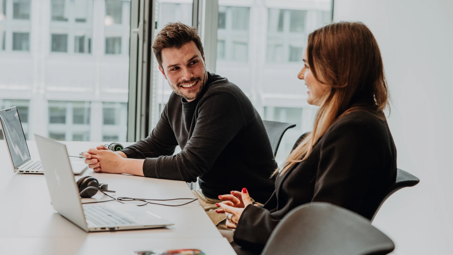 Deux collaborateurs travaillent ensemble sur des ordinateurs portables dans un bureau moderne, échangent avec le sourire et illustrent une atmosphère de travail ouverte et collaborative – aperçu authentique de la culture et du travail chez BSI Software.