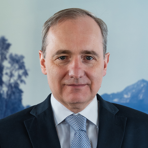 Porträt eines älteren Mannes im Anzug mit blau-weißer Krawatte vor verschwommenem Hintergrund mit Bäumen und Bergen.