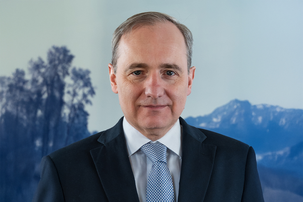 Porträt eines älteren Mannes im Anzug mit blau-weißer Krawatte vor verschwommenem Hintergrund mit Bäumen und Bergen.