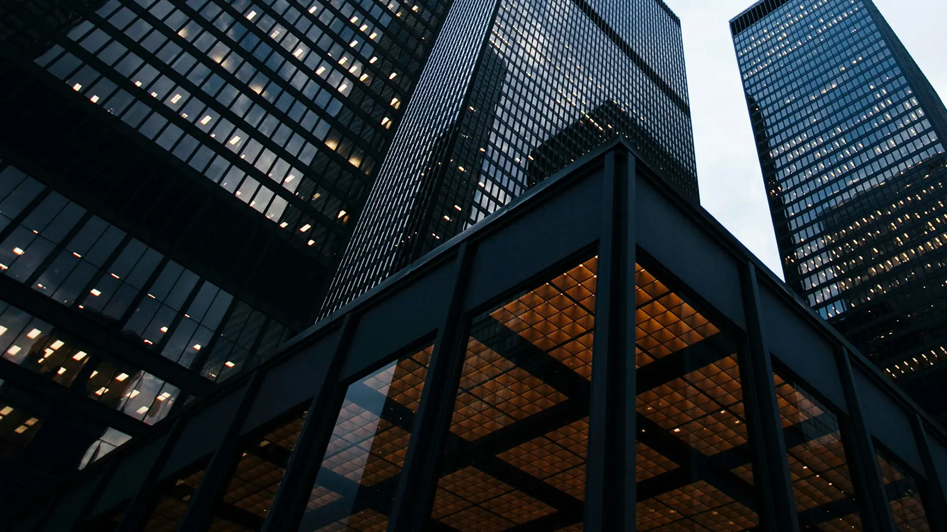 Low-angle view of dark glass bank buildings with illuminated office windows and a striking entrance structure. The modern financial architecture symbolizes PostFinance and Migros Bank expanding their partnership with BSI Software.
