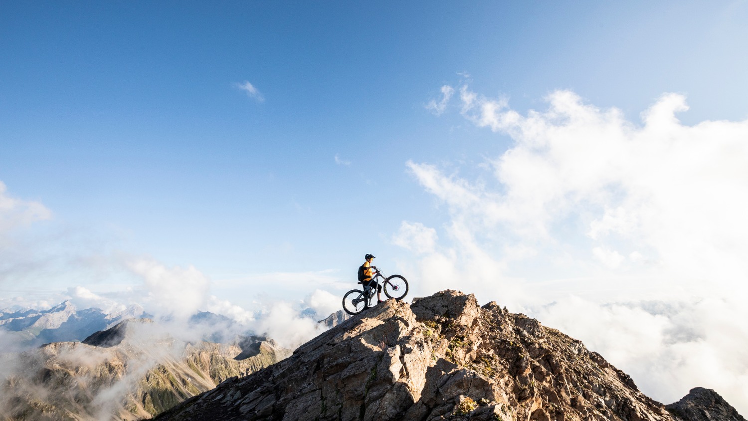 Monika Freiburghaus from BSI Software with a mountain bike on a rocky summit, surrounded by clouds and alpine scenery