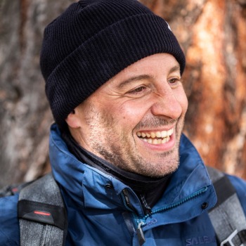 Porträt von Urs Frick, Product Owner CX bei BSI Software, mit Mütze und Outdoor-Jacke, lachend vor einem Baumstamm.