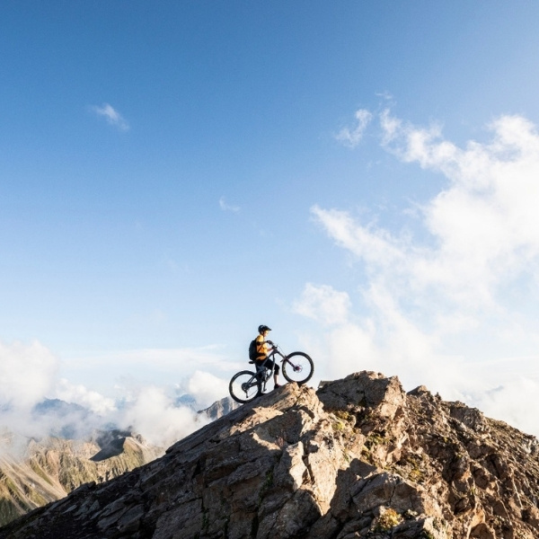 Monika Freiburghaus von BSI Software mit Mountainbike auf einem felsigen Gipfel, umgeben von Wolken und Bergpanorama