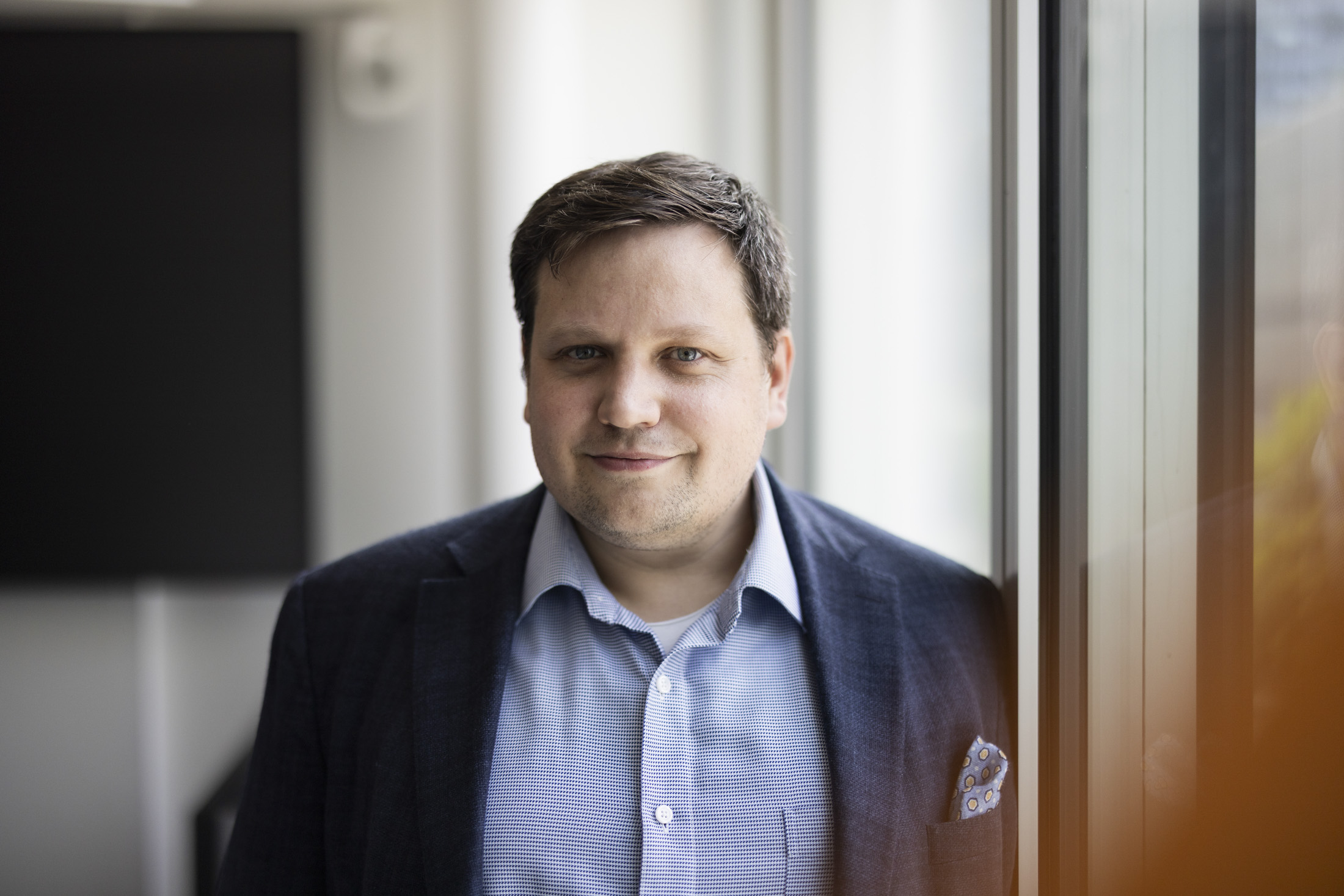 Porträt eines Mannes mit dunklem Sakko und hellblauem Hemd, stehend an einem Fenster mit natürlichem Licht.
