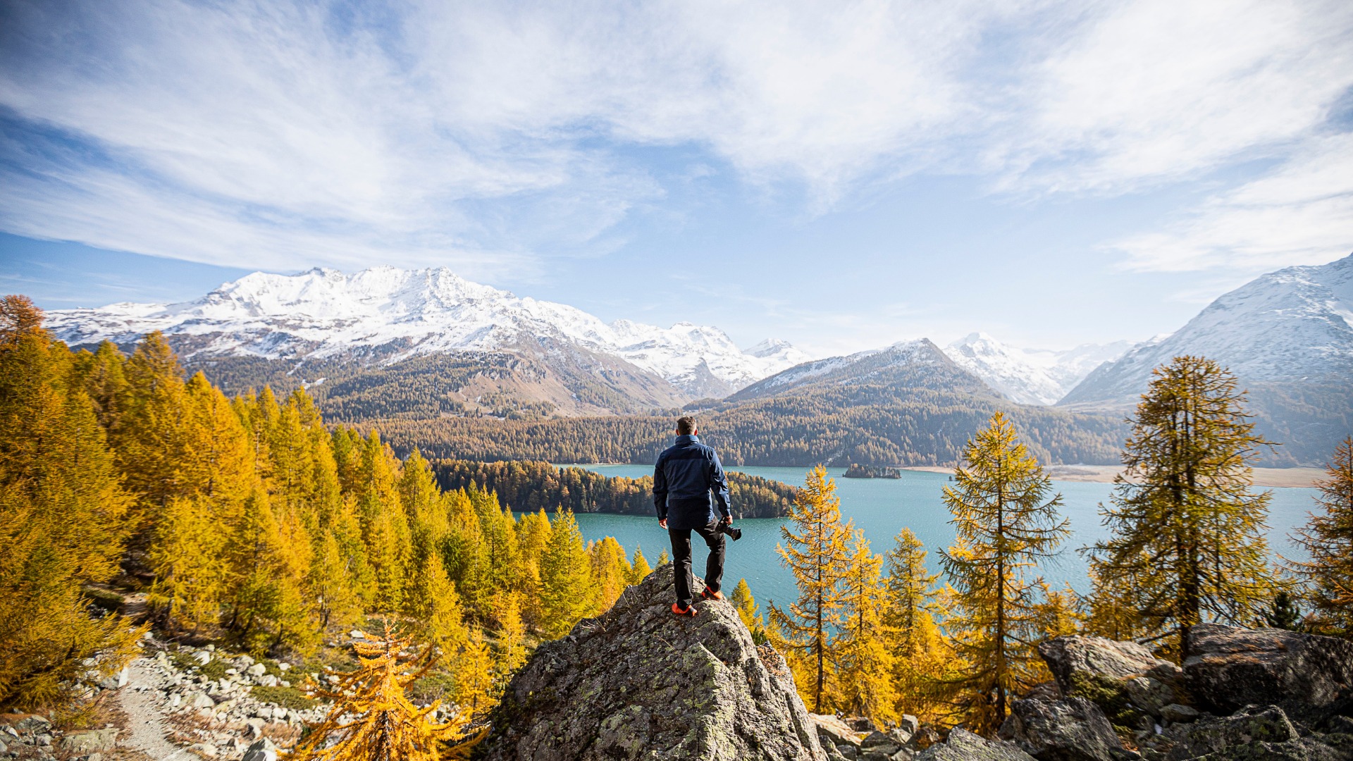 Urs Frick from BSI Software standing on a rock above Lake Sils in the Engadin, holding a camera in his right hand – surrounded by golden larch trees and snow-covered mountains.