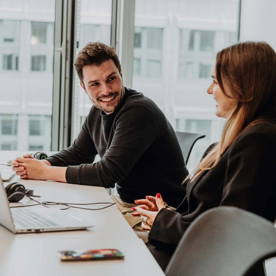 Deux collaborateurs travaillent ensemble sur des ordinateurs portables dans un bureau moderne, échangent avec le sourire et illustrent une atmosphère de travail ouverte et collaborative – aperçu authentique de la culture et du travail chez BSI Software.